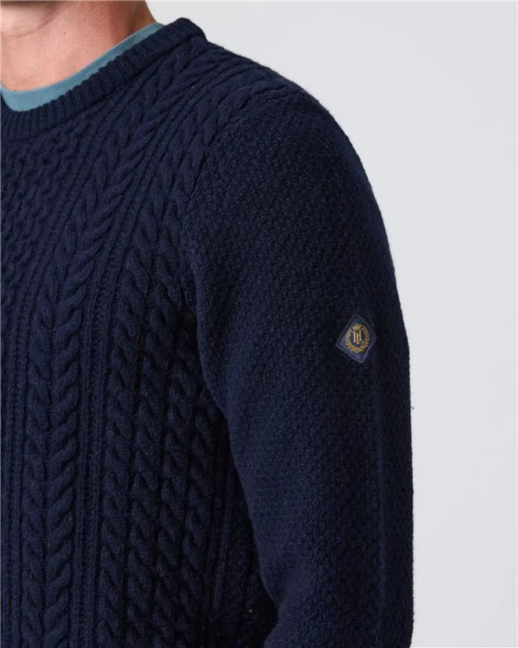 Nærbilde av Henri Lloyd-emblem på Abersoch Cable Knit genser 80 % ull flettestrikk